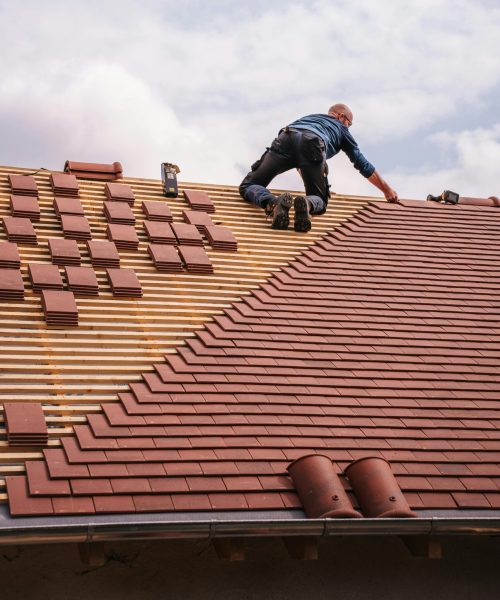 A roofer carefully installs shingles on a new residential roof under a cloudy sky.