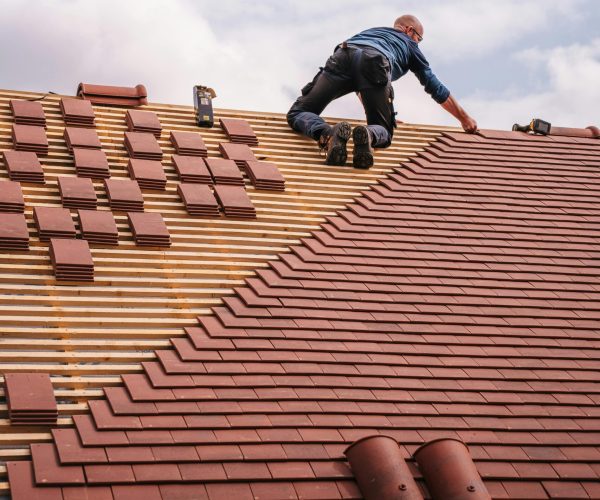 A roofer carefully installs shingles on a new residential roof under a cloudy sky.